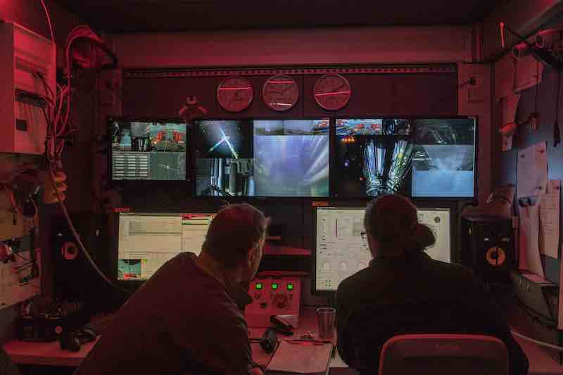 In a small red-lit control room, a man and a woman in front of consoles with several monitors.