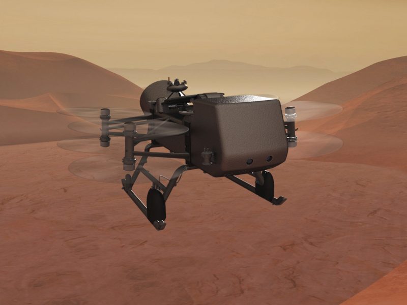 Drone-like machine with four small sets of helicopter blades flying over brownish desert with orangish sky.