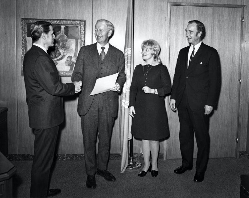 Men in suits and woman in a dress standing in a paneled room with UN flag behind them.