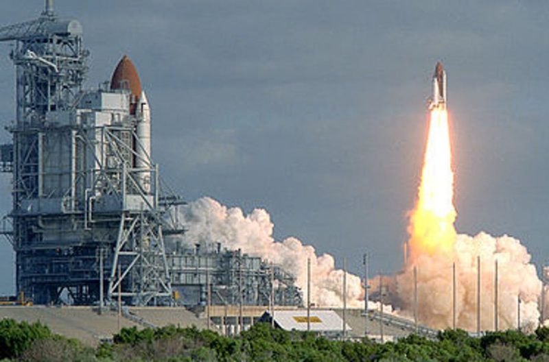 Space shuttle atop a column of flame with billows of steam below, near launch tower with another shuttle.