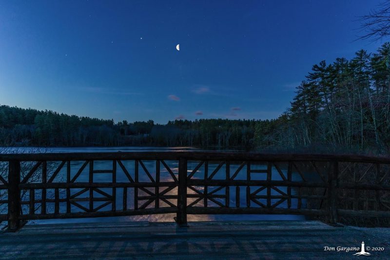 Bridge railing in foreground, moon and planets in background over a lake.