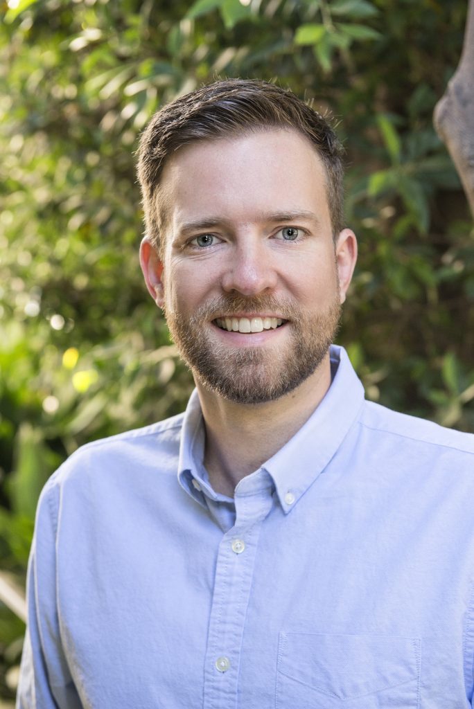 Smiling, bearded young man in dress shirt in front of trees.