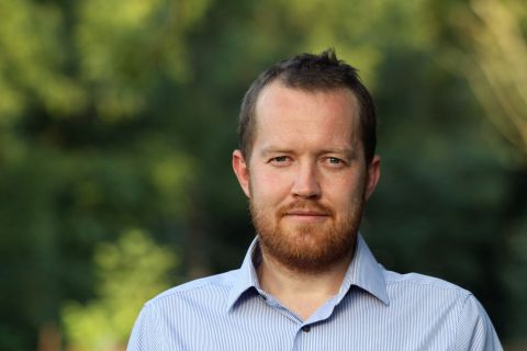 Bearded man in blue dress shirt with trees in background.