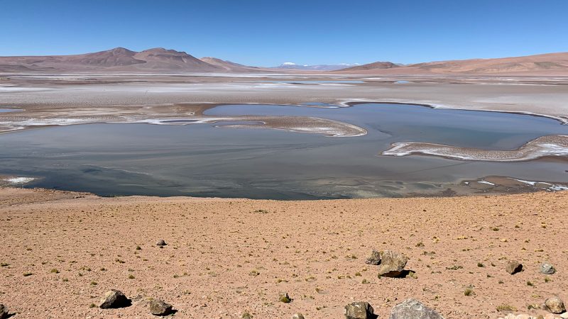 Shallow lake with sandbars and no apparent signs of life around it, with mountains in background under blue sky.