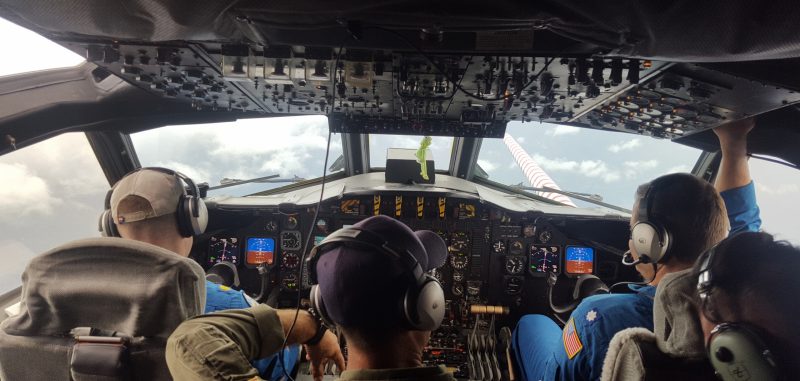 Back view of 4 people in headphones looking toward the front windows of an aircraft.