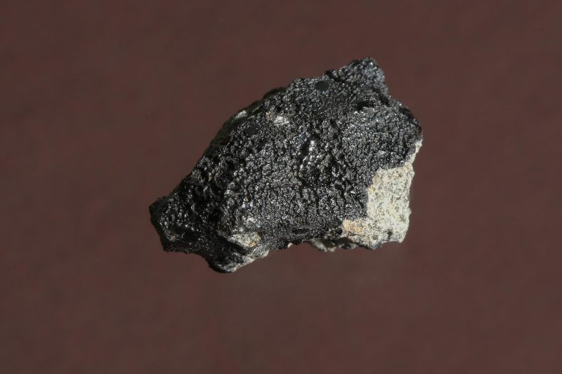 Chunk of dark, irregular, rough-surfaced rock on brownish background.