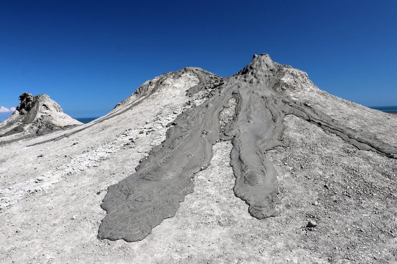 Conical gray colored hill with darker gray semi-liquid-appearing flows on it, with blue sky in background.