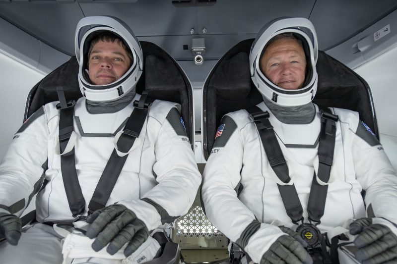 Two men in white space suits with the faceplates open, strapped into seats in a spacecraft.