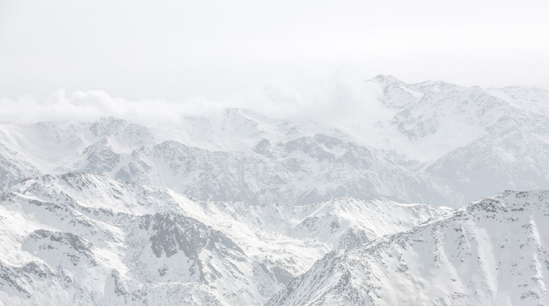 Snow-covered mountain peaks seen from above going into distant hazy horizon under white sky.