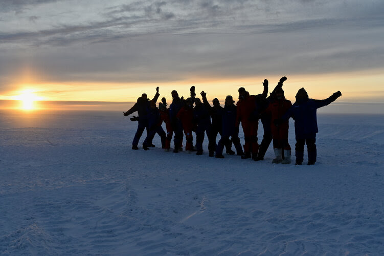 A line of about ten people standing in snow, waving and posing, silhouetted against the sun setting behind them.