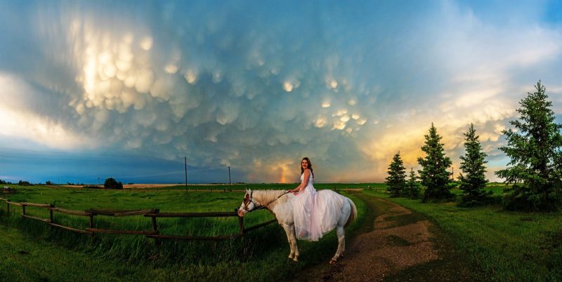 Young woman in long white dress on white horse under sky with many small, sunlit downward bulges from a dark cloud.