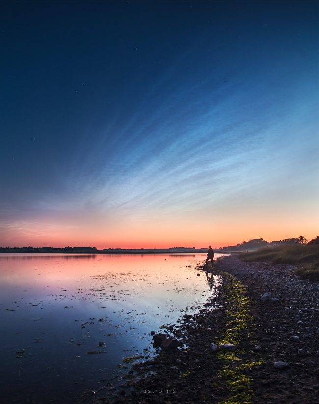 High, rippled, shining clouds above waterway in twilight, man standing on beach.
