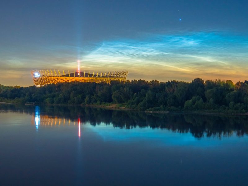 Electric blue noctilucent clouds shining above a river with a lit-up stadium in the distance.