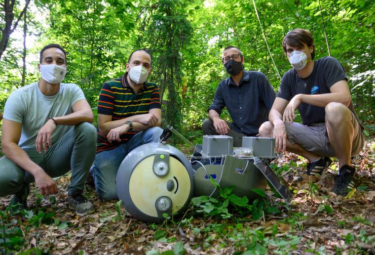 Four men in masks and casual garb squatting next to robot on the ground. Robot has sloth-like face.