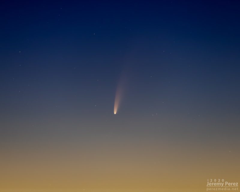 A bright spot, the comet, with long wedge-shaped tail upward in a sunset sky.