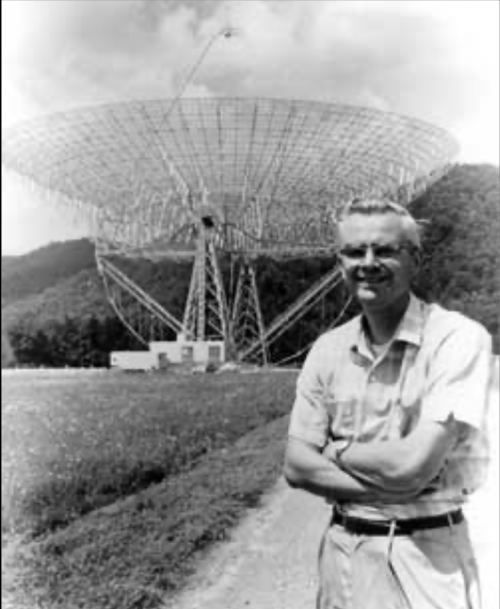 Young scientist, arms folded, standing in front of dish-type radio telescope.