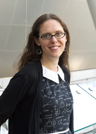 Smiling, long-haired woman with glasses and blouse covered with mathematical symbols.