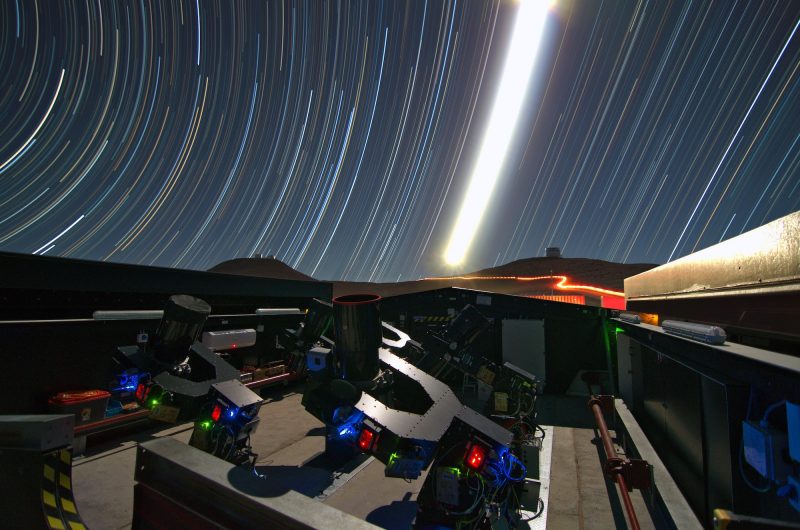 Telescopes on ground with long, bright, concentric curved lines in the sky.