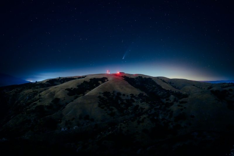 Um cometa no céu, com vermelho iluminado construção no topo de uma colina em primeiro plano, e uma noite de brilho das luzes da cidade ao fundo.