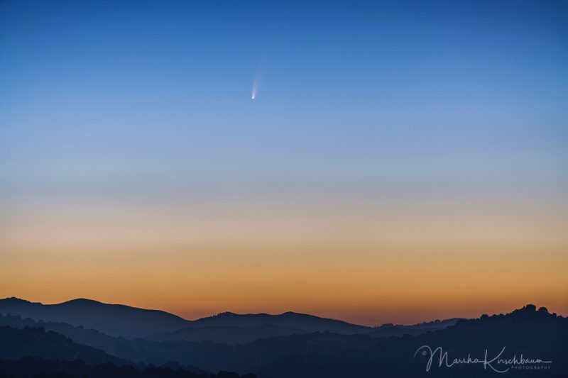 Un cometa con una cola dividida contra un cielo crepuscular brillante sobre colinas azules.