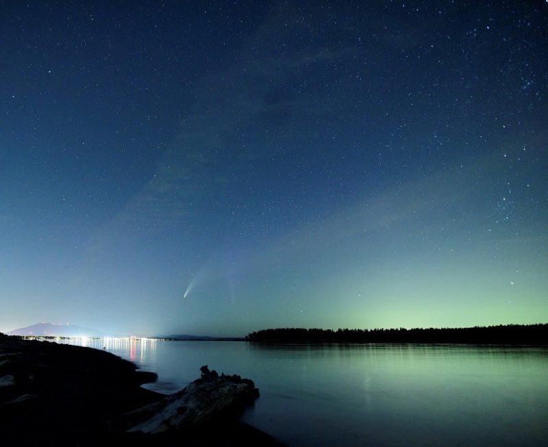 komeet en aurora tegen een medium blauwe sterrenhemel, over een waterlichaam.