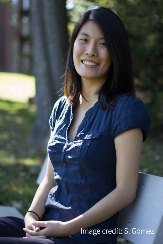 Smiling woman in blue blouse, sitting on bench with tree in background.