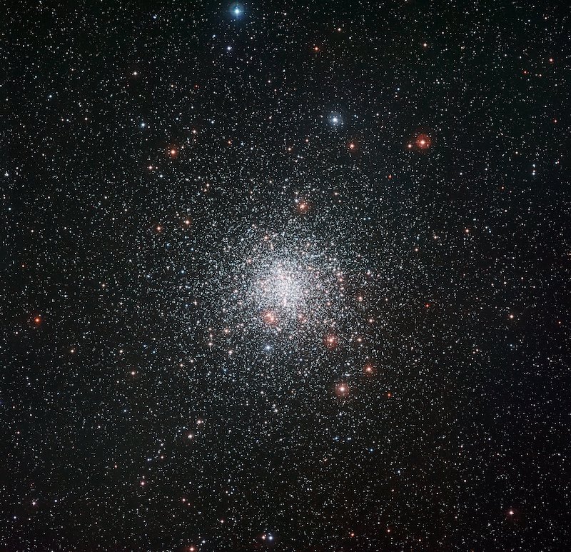 A tight cluster of faint stars, mostly white, in a dense star field with some yellow and red stars.