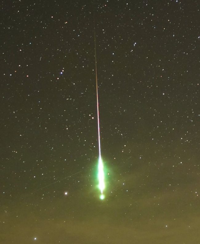 Bright elongated streak with large greenish head pointing downward, in starry sky.