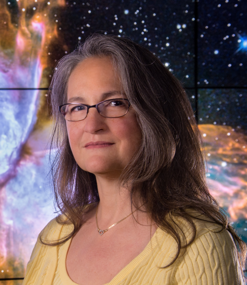 Woman with long hair, glasses and yellow blouse, with nebula mural in background.