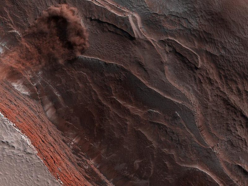 Nuage poussiéreux à la base d'une falaise abrupte dans un terrain rougeâtre.