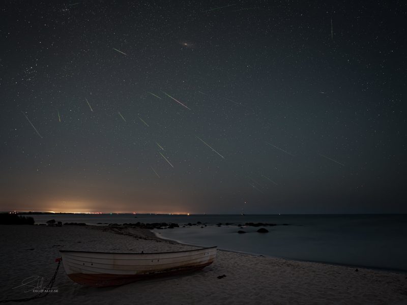 Many thin bright streaks in the sky over shoreline with small boat pulled up on the sand.