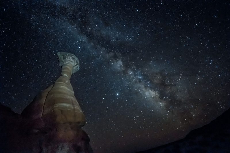 Looking upward along a tall cylindrical rock formation at the Milky Way crossing a dark sky.