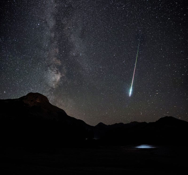 Observing meteors over dark hills, with Milky Way and a thin white streak bigger at lower end.