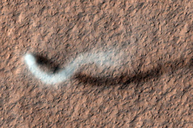 A white S-shaped plume over reddish rocky terrain as seen from orbit.
