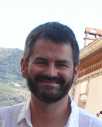 Smiling man with moustache and beard, with building and hill behind him.