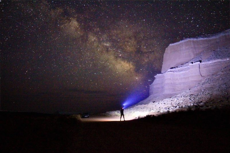 Man with headlamp illuminating rock cliff, Milky Way in background.