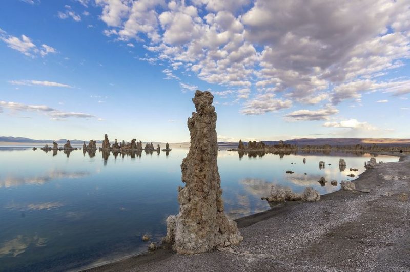 Lake in bleak landscape with tall, rough pillars around shoreline and blue sky with clouds.