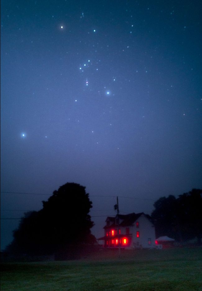 Orion and Sirius in misty blue sky above old two-story farmhouse with lighted windows.