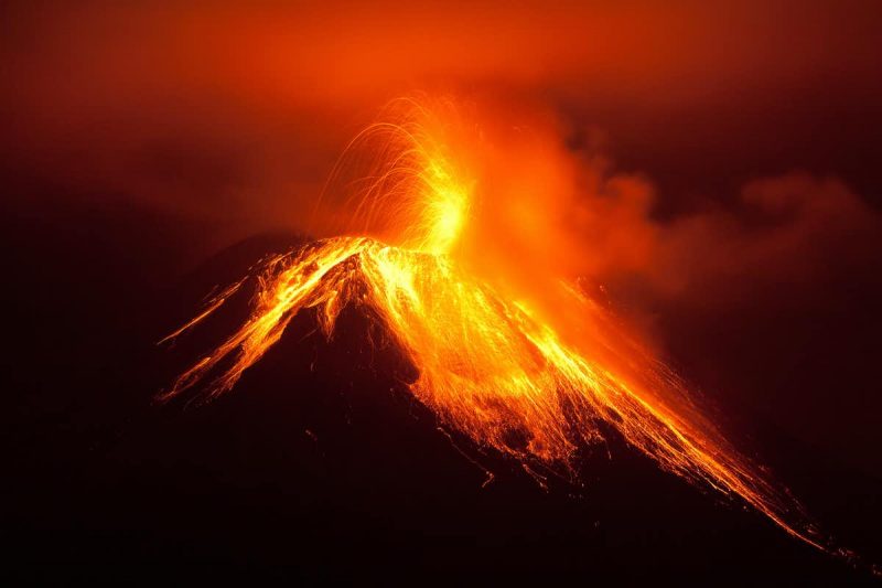 Volcano at night with glowing lava pouring down its sides and explosions at top.