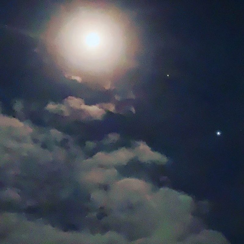 Bright over-exposed moon casting light on clouds, with Jupiter and Saturn nearby.