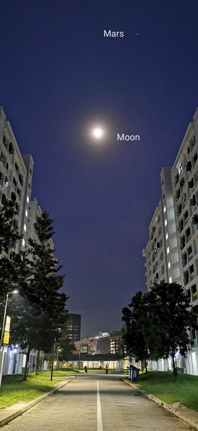 The glowing moon and Mars above buildings along a street in Singapore.