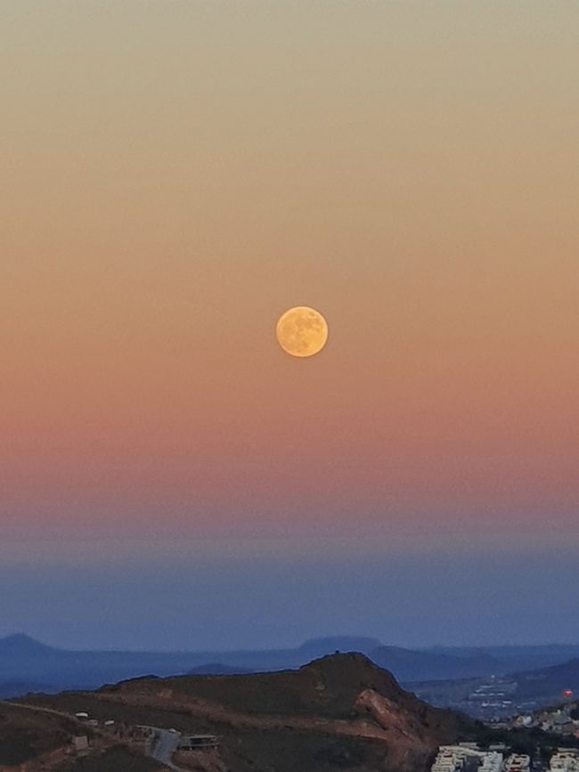 Yellow moon floating in pink sunset sky over blue hills.