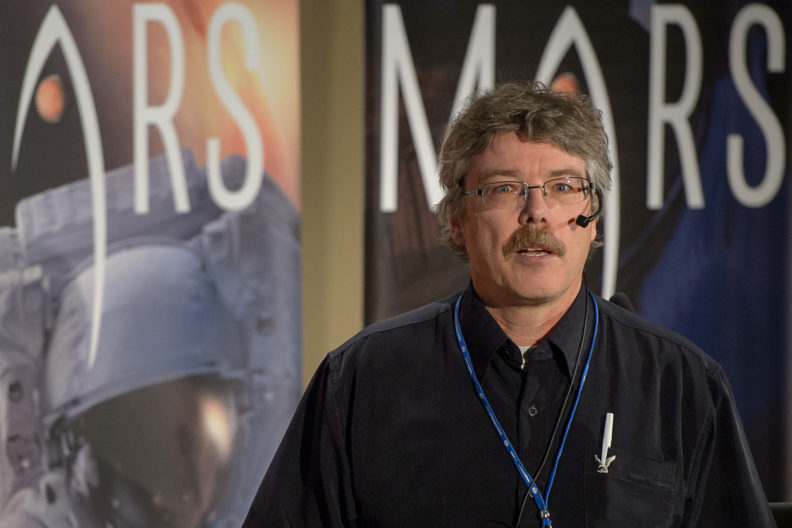Man with microphone and blue lanyard over black shirt, with the word Mars behind him in large letters.