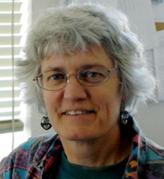 Smiling gray-haired woman with eyeglasses, with window blinds behind her.