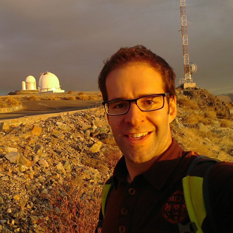 Smiling man with eyeglasses; visible in distance, telescope domes and radio tower.