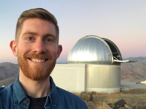 Smiling, bearded, young man standing in front of an open telescope dome.