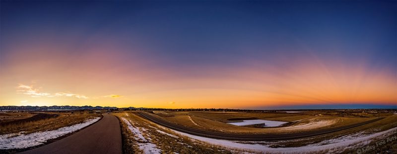 Anticrepuscular rays: Panorama of mostly barren landscape and road with orange glow and higher dark blue sky.