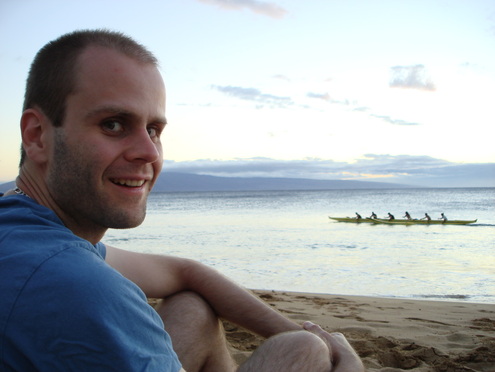 Smiling, short-haired man in blue shirt with kayak on lake in the distance.