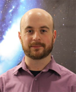 Balding young man with beard, standing in front of an image of a galaxy.