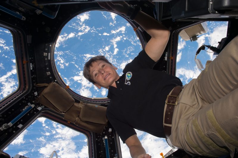 Woman floating in multi-windowed chamber of ISS with blue and white Earth visible through windows.